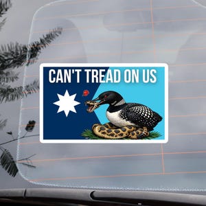 May include: A rectangular sticker with the text "CAN'T TREAD ON US" above a design featuring a loon attacking a rattlesnake. The design includes a white star on a blue background and a red berry. The sticker is on a car window.