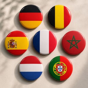 May include: Seven circular pin-back buttons featuring national flags. The flags displayed are Germany, Belgium, Spain with its coat of arms, France, Morocco with a green star, the Netherlands, and Portugal with its coat of arms, all arranged in a group.