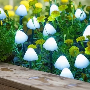 May include: A garden bed features several white mushroom-shaped lights among green foliage and yellow flowers. The lights are illuminated, creating a soft glow. The scene is set in a wooden planter box, suggesting outdoor decor.