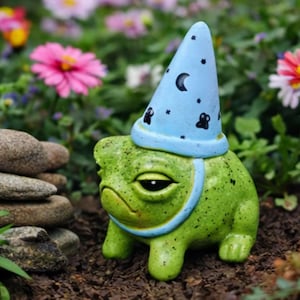 May include: A quirky green frog ornament wearing a light blue wizard's hat decorated with black stars and a crescent moon. The frog has a grumpy expression, set in a garden.