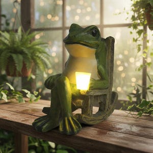 Peut inclure: Une statue de grenouille verte assise dans un rocking-chair en bois, tenant une lumière carrée brillante. La grenouille a un ventre jaune clair et est posée sur une surface en bois. L'arrière-plan comprend une fenêtre et des plantes suspendues.