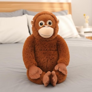 以下が含まれることがあります： 茶色のぬいぐるみオランウータン。明るいタン色の顔と大きな目があります。ぬいぐるみは直立しており、腕と脚が伸びています。本物のオランウータンに似せてデザインされています。