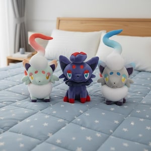 Puede incluir: Tres peluches con forma de criaturas estilizadas parecidas a gatos. Los juguetes son en tonos de azul, verde y gris, con detalles en rojo, amarillo y blanco. Tienen orejas grandes y apéndices largos y curvos. Sobre un edredón azul.