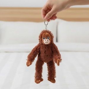 Może przedstawiać: Brązowy pluszowy brelok do kluczy z orangutanem z uśmiechniętą twarzą i metalowym kółkiem. Pluszowa zabawka ma długie ramiona i nogi i jest wykonana z miękkiego, puszystego materiału. Tło to łóżko z białymi prześcieradłami.