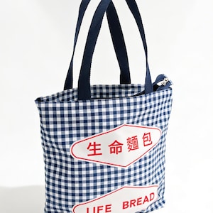 Puede incluir: Un bolso tote a cuadros azules y blancos con asas azul marino. La bolsa presenta dos etiquetas blancas y rojas con el texto "LIFE BREAD" y caracteres chinos. La bolsa tiene un cierre de cremallera.