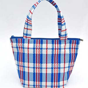 Puede incluir: Un bolso tote con cremallera y asas azules. El bolso presenta un fondo blanco con un patrón de cuadros de líneas verticales y horizontales rojas y azules. Las asas y la cremallera son azules.