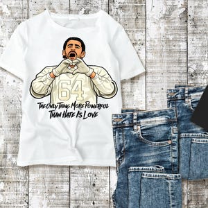 May include: White t-shirt with a graphic of a person forming a heart shape with their hands. The shirt displays the number 64 and the text "THE ONLY THING MORE POWERFUL THAN HATE IS LOVE". Folded blue jeans are placed beside the shirt.