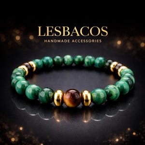 Puede incluir: Una pulsera de cuentas con esferas verdes estilo malaquita, dos cuentas marrones de ojo de tigre y espaciadores de metal dorado brillante. El texto LESBACOS HANDMADE ACCESSORIES aparece sobre la pulsera en un fondo oscuro.