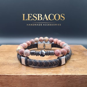 Può includere: Due braccialetti su una superficie di legno. Il braccialetto superiore presenta perline di pietra marrone-rossastro. Il braccialetto inferiore ha un cinturino in pelle marrone scuro intrecciato con dettagli metallici argentati. Il marchio "LESBACOS" e il testo "HANDMADE ACCESSORIES" sono sullo sfondo.