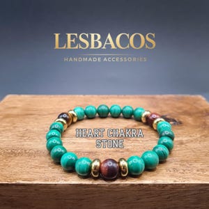 May include: A handmade bracelet featuring green malachite beads, interspersed with gold-colored spacers and brown beads. The text "HEART CHAKRA STONE" is displayed on the wooden surface. The brand name "LESBACOS" and "HANDMADE ACCESSORIES" are in the background.