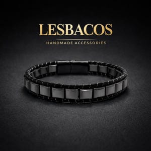 Può includere: Un bracciale nero caratterizzato da una fila di perline metalliche quadrate dai toni scuri, integrate in un cinturino in corda intrecciata con una chiusura rettangolare liscia. Sopra, il testo LESBACOS HANDMADE ACCESSORIES è in lettere dorate.