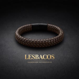 Puede incluir: Una pulsera de cuero trenzado marrón con un cierre negro. La pulsera tiene forma circular y se muestra sobre un fondo oscuro. El nombre de la marca "LESBACOS" y "HANDMADE ACCESSORIES" están impresos en dorado.
