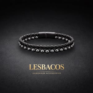 Op de afbeelding: Een dubbelstrengs armband met een gevlochten zwart leren koord en een streng gepolijste donker-metalen kralen. De armband is voorzien van een matzwarte cilindrische magnetische sluiting. De tekst LESBACOS HANDMADE ACCESSORIES staat eronder afgebeeld.
