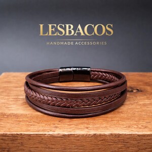 Pode incluir: Uma pulseira de couro castanho de várias camadas com cordões de couro liso e um cordão central entrançado. A pulseira é fixada com um fecho magnético de metal preto. O texto LESBACOS HANDMADE ACCESSORIES é exibido acima.