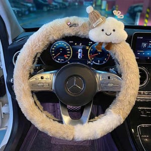 Peut inclure: Un couvre-volant beige et duveteux avec une peluche en forme de nuage et un petit bonnet en tricot. Le couvre-volant est installé sur le volant d'une voiture, avec le tableau de bord et les jauges visibles en arrière-plan. Le mot "mua" est imprimé sur le couvre-volant.