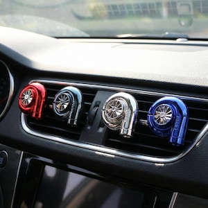 Könnte beinhalten: Vier Auto-Lufterfrischer in Form von Turboladern in Rot, Blau, Silber und Dunkelblau. Die Lufterfrischer sind an einer Lüftungsöffnung im Auto befestigt. Das Wort "TURBO" ist auf dem blauen Lufterfrischer zu sehen.