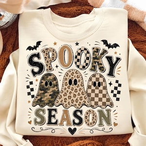 Op de afbeelding: Een crèmekleurig sweatshirt met ronde hals en de tekst SPOOKY SEASON in grote, gedessineerde letters. Onder de tekst staan drie spookillustraties met camouflage-, luipaard- en dambordpatronen, geaccentueerd door vleermuizen en sterren.