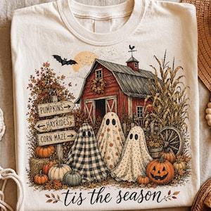 Può includere: Una t-shirt color crema con una grafica raffigurante un fienile rosso, tre fantasmi, zucche e un cartello con il testo PUMPKINS, HAYRIDES e CORN MAZE. Il design include un pipistrello, una luna e la scritta tis the season in basso.