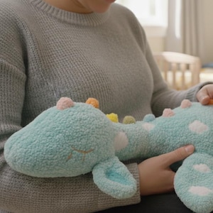 Könnte beinhalten: Ein weiches, hellblaues Plüsch-Dinosaurier mit weißen Punkten und bunten Akzenten. Das Stofftier hat geschlossene Augen und wird gehalten. Das Plüschtier ist eine niedliche Ergänzung für ein Kinderzimmer.