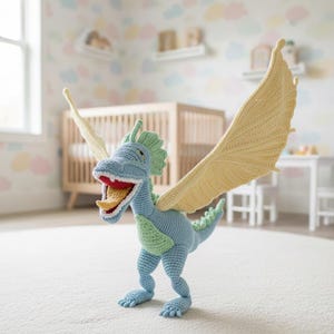 Puede incluir: Un juguete de dragón de ganchillo hecho a mano en tonos de azul, verde y amarillo. El dragón tiene alas extendidas, una boca abierta con interior rojo y lengua amarilla, y está de pie. El juguete es para niños.