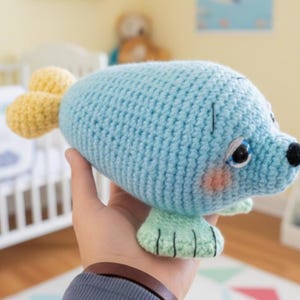 May include: A light blue crocheted seal plush toy with yellow tail fins and light green flippers. The seal has a black nose, blue eyes, and pink cheeks. The handmade toy is designed for children.