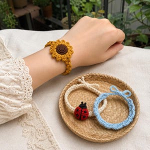 May include: A set of three crochet hair ties featuring a yellow sunflower, a cream-colored band with a red and black ladybug, and a light blue band with a small bow. Each accessory is crafted from yarn with distinct floral and insect motifs.