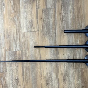 May include: Three black telescoping flag poles of varying lengths, each with a circular base. The poles are lying on a light brown wooden surface. The poles are fully extended.