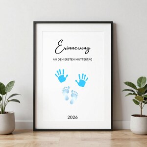 May include: Framed print with the German word "Erinnerung" and the phrase "AN DEN ERSTEN MUTTERTAG". Features blue handprints, footprints, and the year 2026. The print is in a black frame.