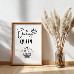 May include: A framed print with the words "Baking QUEEN" in black script and a cupcake illustration. The frame is light brown, and the print is displayed on a wooden surface. A vase of dried pampas grass is next to the frame.