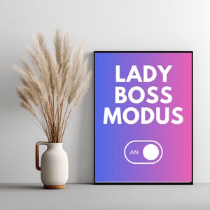 May include: A framed print with the words "LADY BOSS MODUS" in white letters against a blue and pink gradient background. A white "ON" switch graphic is below the text. A vase with dried pampas grass is to the left.