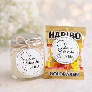 Puede incluir: Un tarro de cristal con tapa plateada y lazo de cuerda, y una bolsa dorada de gominolas HARIBO. Ambos artículos tienen una pegatina redonda blanca con el texto "Schön, dass du da bist" y un corazón. El tarro está lleno de una sustancia blanca.