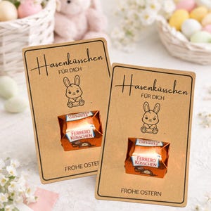 Può includere: Due biglietti marroni con la scritta "Hasenküsschen FÜR DICH" e l'illustrazione di un coniglietto dei cartoni animati. Ogni biglietto contiene un cioccolatino Ferrero Rocher incartato. Le parole "FROHE OSTERN" sono stampate in basso su ogni biglietto. A tema pasquale.