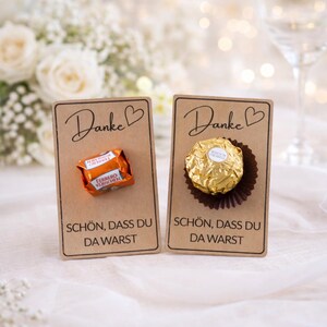 Puede incluir: Dos tarjetas marrones pequeñas con la palabra "Danke" y un corazón, y el texto "SCHÖN, DASS DU DA WARST". Una tarjeta tiene un chocolate envuelto en naranja, la otra en oro. Flores blancas borrosas en el fondo.
