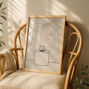 Könnte beinhalten: Ein gerahmter Kunstdruck mit einer einfachen schwarzen Strichzeichnung einer kleinen Katze, die hinter einer Toilettenpapierrolle hervorschaut. Die Illustration ist auf einem hellbeigen Hintergrund zentriert und in einem hellen Holzrahmen.