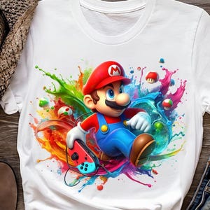 Könnte beinhalten: Weißes T-Shirt mit einer farbenfrohen Grafik von Mario, der Videospiel-Figur, die einen Controller hält. Mario trägt seinen roten Hut, blaue Latzhose und braune Schuhe, umgeben von lebendigen Farbspritzern. Das Design beinhaltet Pilze und andere Spielelemente.