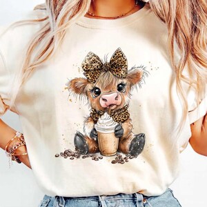 Könnte beinhalten: Cremefarbenes T-Shirt mit einem Cartoon-Kalb mit Leopardenmuster-Schleife und Schal, das einen Kaffeebecher hält. Das Kalb ist braun mit großen Augen, umgeben von Kaffeebohnen. Das T-Shirt ist ein lässiger Stil.