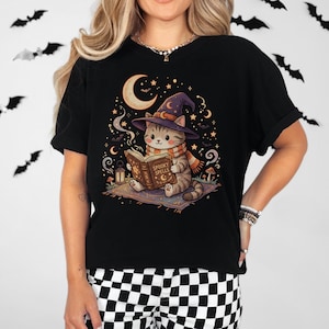 Puede incluir: Una camiseta negra con el gráfico de un gato atigrado que lleva un sombrero de bruja morado y una bufanda naranja, leyendo un libro titulado Spooky Spells. La ilustración incluye una luna creciente, estrellas, murciélagos, una linterna y setas.
