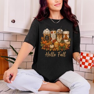 Puede incluir: Una camiseta negra con un estampado gráfico de cuatro vasos de café helado, calabazas, hojas de otoño y una vela. El texto "Sippin on Magic", "Hello Fall" y "Harvest Spice" está impreso en los vasos y la vela. Las palabras "Hello Fall" aparecen en letra dorada debajo del gráfico.
