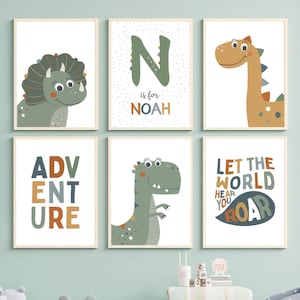 May include: Six framed dinosaur-themed art prints. The prints feature cartoon dinosaurs, the word "ADVENTURE", the letter "N" with the name "NOAH", and the phrase "LET THE WORLD HEAR YOU ROAR". The color palette includes green, orange, and blue.