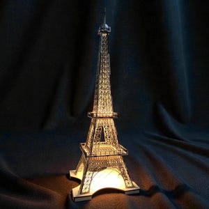 Puede incluir: Una detallada maqueta metálica de la Torre Eiffel con una fuente de luz interna, que emite un brillo cálido. El intrincado diseño de celosía es visible, y la torre se alza sobre una base cuadrada. El fondo es una tela oscura y texturizada.