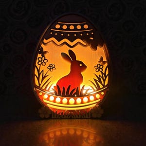Può includere: Una luce notturna in legno a forma di uovo a tema pasquale. L'interno presenta una silhouette di coniglio in un campo con fiori e farfalle. La luce emette una luce arancione. La base ha un design floreale decorativo. Misura circa 15 cm di altezza.