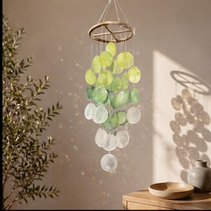 May include: A hanging wind chime with a wooden ring and strands of translucent, round, green and white shells. The shells graduate in color from light green to white. The wind chime hangs in front of a neutral-colored wall.