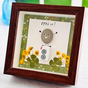 May include: A dark wood shadow box frame containing a pebble art figure with a smiling face and raised arms. The figure stands on a white mahjong tile marked with the number 2 and two circular symbols. The background features yellow flowers.