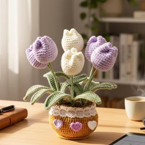 May include: A crochet tulip display in a decorative pot. The flowers are in shades of purple and white, with green leaves. The pot is yellow with a lace trim and heart accents. A charming, handmade home decor item.