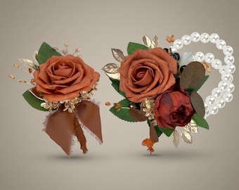 May include: A boutonniere and a corsage featuring burnt orange roses, dark red accents, green leaves, and gold-toned foliage. The corsage includes a double-strand white pearl bracelet, while the boutonniere is finished with a brown ribbon.