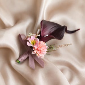 May include: A boutonniere featuring a deep dark purple calla lily, a small light pink flower with a yellow center, and a cluster of soft pink petals. The stems are wrapped in a dusty mauve ribbon that forms a small bow at the base of the flowers.