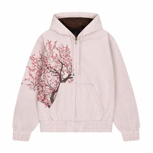 May include: A light pink zip-up hooded jacket with a brown interior lining. The jacket features a detailed illustration of a cherry blossom tree on the left side. The jacket has a drawstring hood and two front pockets.