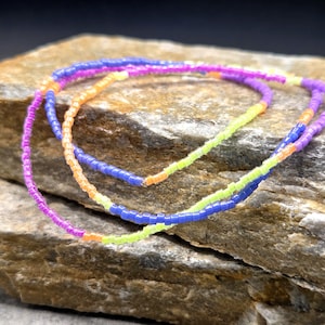 May include: Three beaded necklaces in vibrant colors: purple, orange, blue, and neon green. The necklaces are made of small, colorful beads and are arranged in a layered style. The necklaces are displayed on a textured, light brown stone.