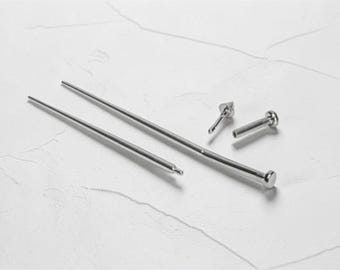 May include: A collection of silver-colored metal hair pins and accessories. The image shows several straight pins with rounded and decorative ends, along with a few smaller, decorative pieces. The items are arranged on a white textured surface.