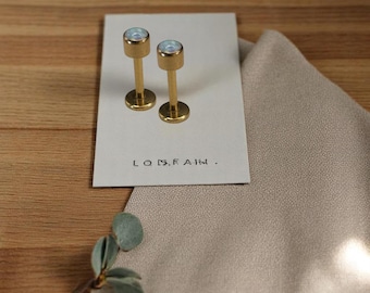 May include: Two gold-toned metal stud earrings with iridescent circular tops, displayed on a rectangular white card featuring the printed text "LO F A". The earrings have a cylindrical post design and are positioned side-by-side on the card.